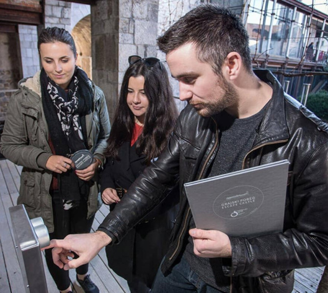 Trije ljudje stojijo pred stojalom s tipikami, v ozadju prostori gradu.