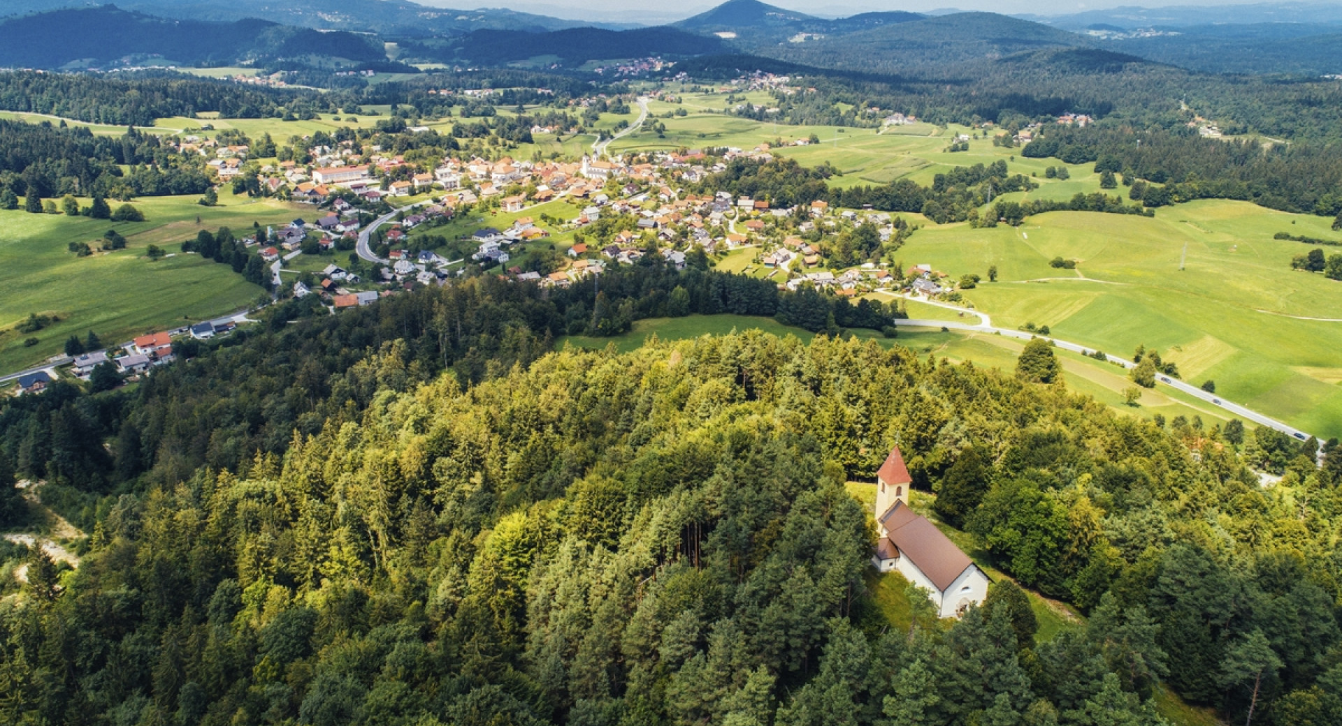 Kraj, ki ga obdaja zelena pokrajina z gozdovi, sredi katerih je tudi cerkev.