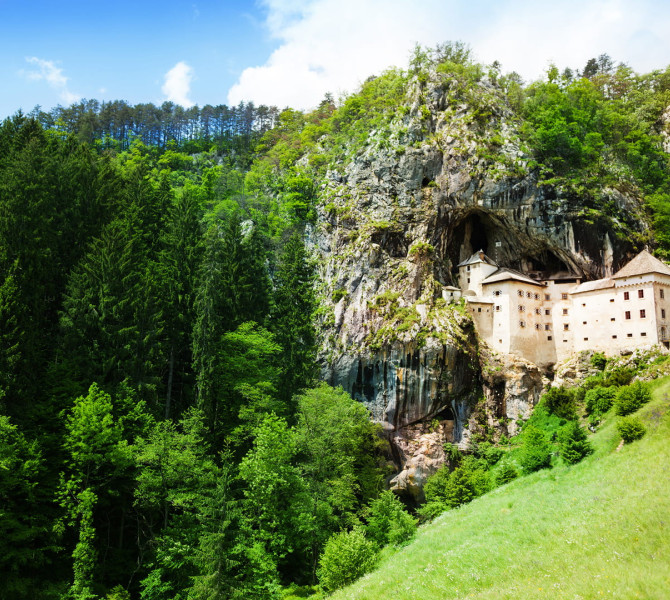 Il castello medievale di Predjama incastonato in una grotta rocciosa, circondato da boschi e prati sotto un cielo azzurro.