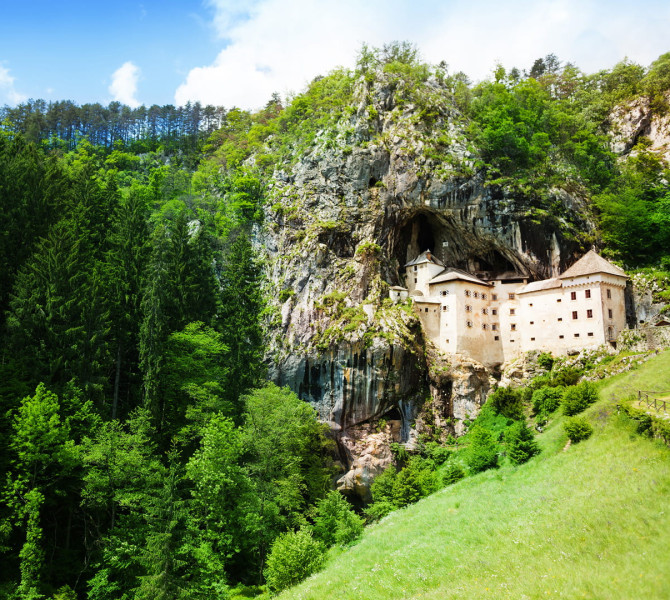 Predjama medieval castle built into a rocky cave, surrounded by forest and meadows under a blue sky.