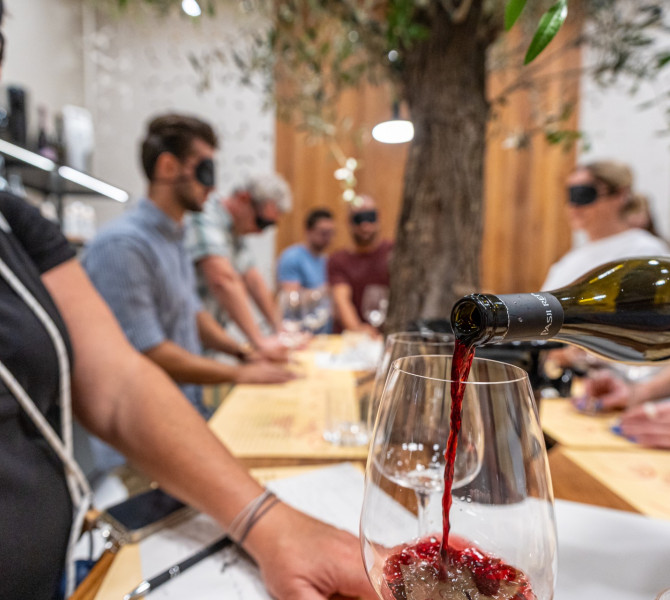 Un primo piano del versare vino rosso in un bicchiere, con delle persone che aspettano in fila sullo sfondo.