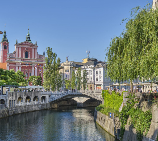 Reka skozi mesto, na nabrezjih zelenje in v ozadju rdeca cerkev, most in mescanske hise.
