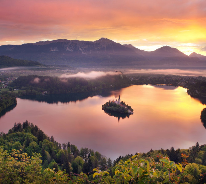 A lake with an island, surrounded by forest and hills at sunset.