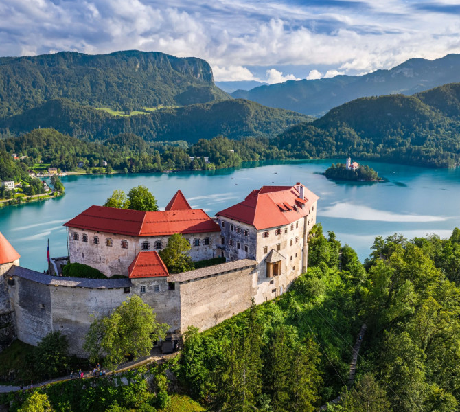 A castle on a hill, below it a lake with an island, surrounded by forest.
