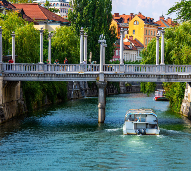  Most cez reko, po reki pluje ladjica, v ozadju zelenje in mescanske hise.