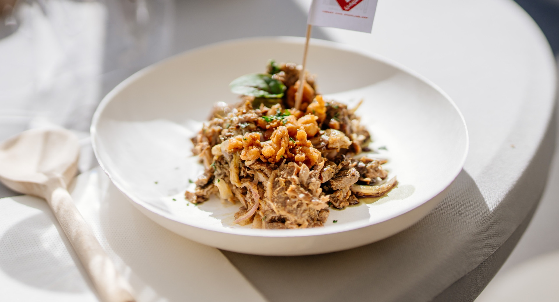 A white plate containing a Slovenian pulled pork or beef dish, topped with cracklings and herbs, decorated with a small flag, and served on a table next to a wooden spoon.