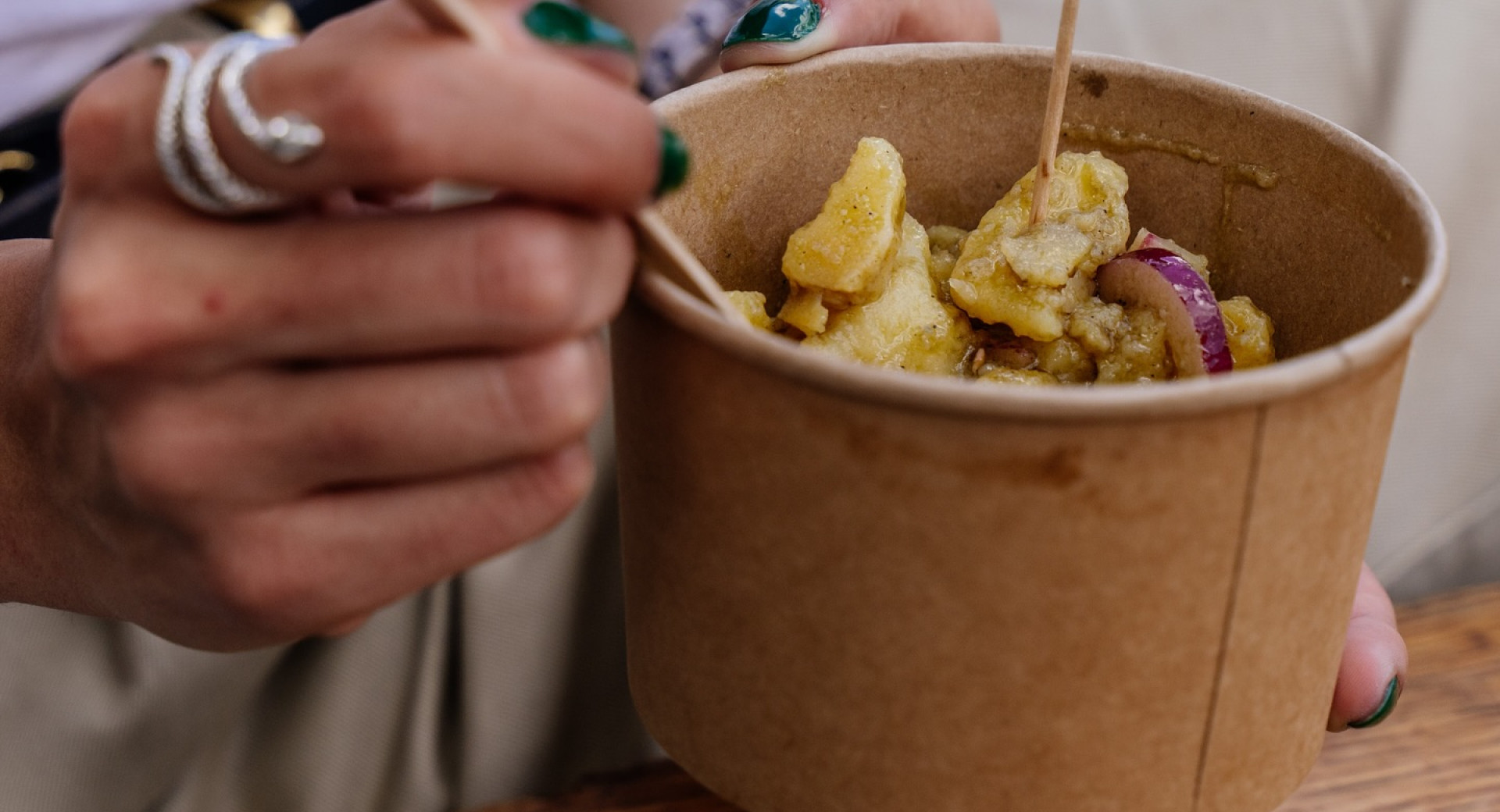 Nahaufnahme einer Person mit Ringen und grünen Fingernägeln, die hausgemachten Kartoffelsalat mit roten Zwiebeln mit einem Holzlöffel aus einer Pappschale isst.
