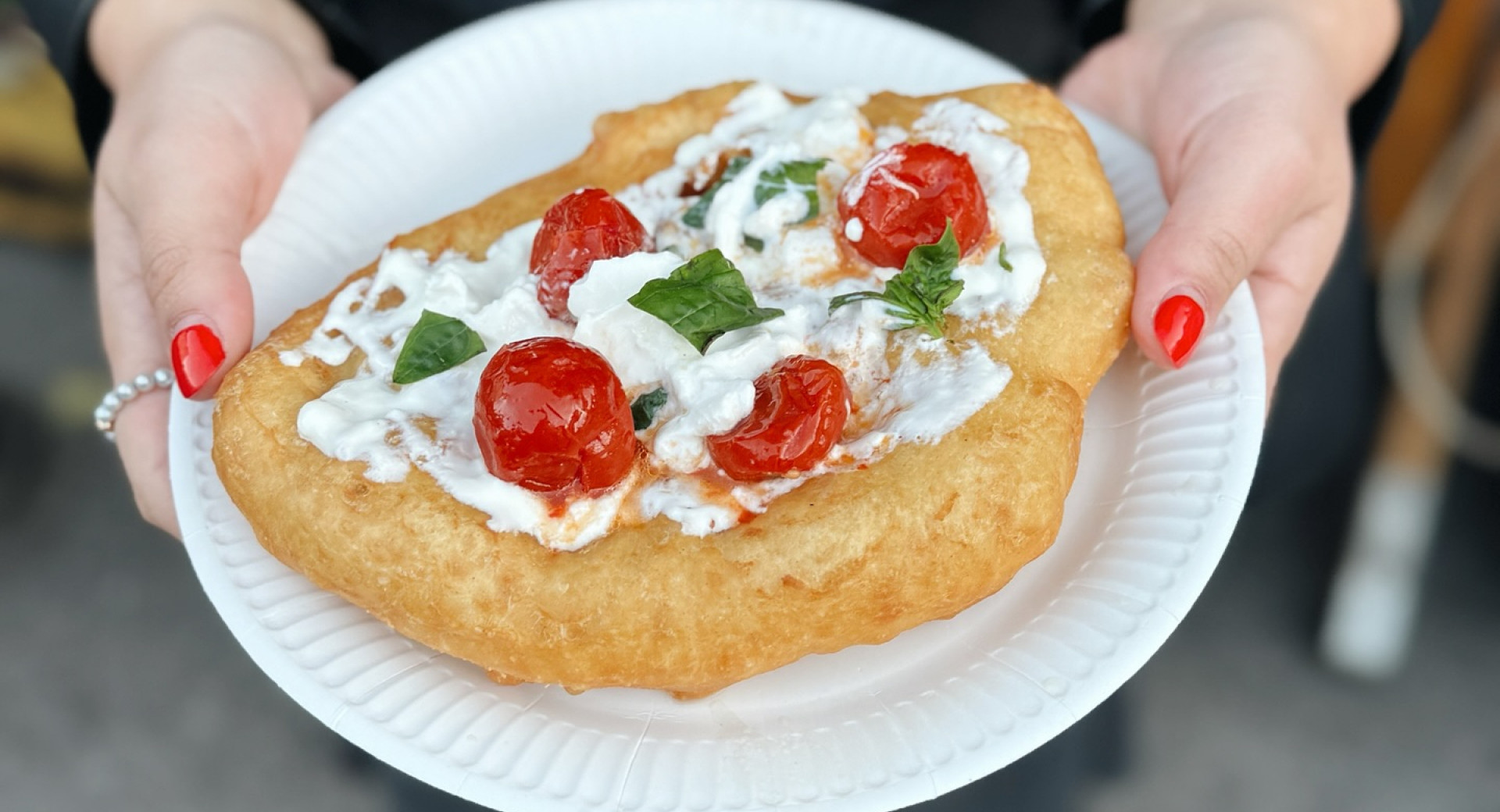 Eine Person mit roten Fingernägeln und einem Perlenring hält einen weißen Teller mit goldbraun frittiertem Teig, belegt mit frischer Burrata, gerösteten Kirschtomaten und Basilikumblättern.