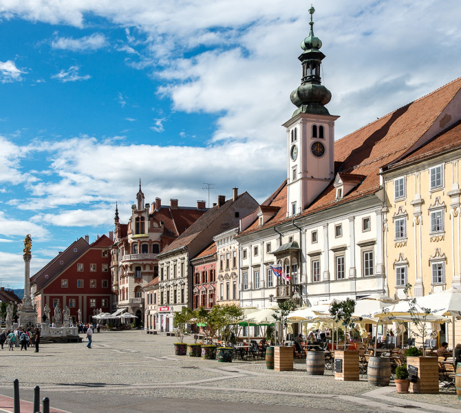 A town square with a church and bourgeois houses on the right; tables, chairs, and white parasols in front of one house.