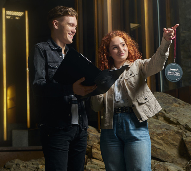 Fant v rokah drzi crno mapo z navodili, dekle pa crni obesek z logom Ljubljanskega gardu.