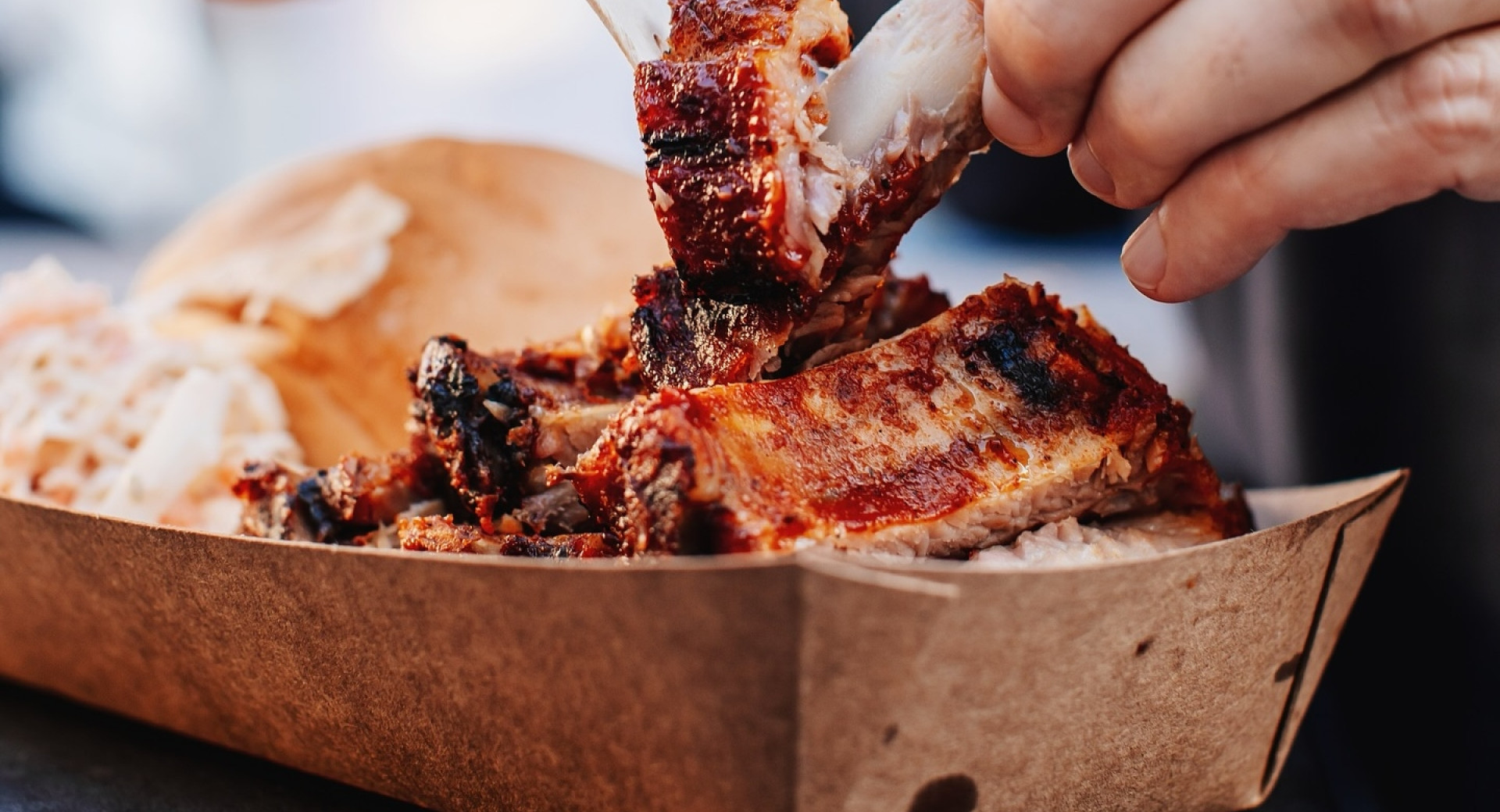 Eine Person zerreißt mit Händen und einer Holzgabel ein Stück saftige, angekohlte BBQ-Rippchen, die in einer Papierverpackung bei einem Street-Food-Event serviert werden.