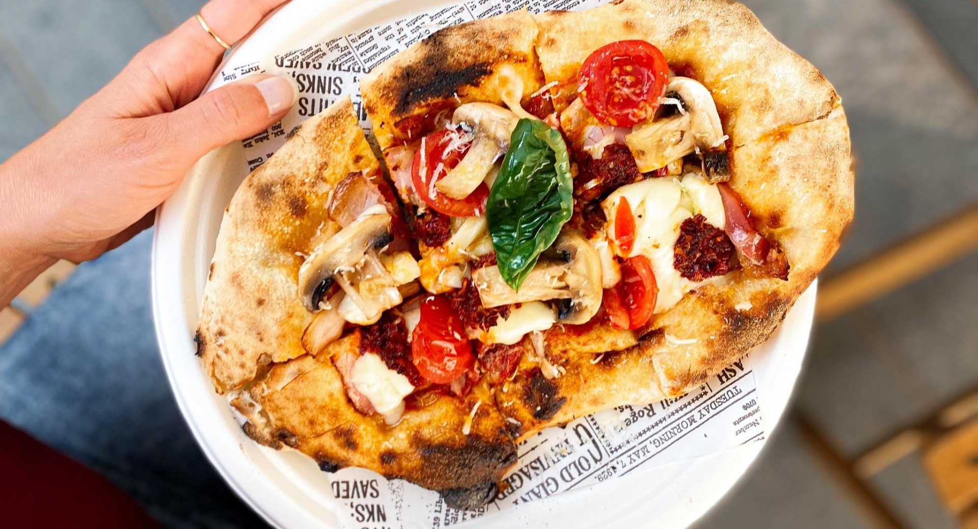 Eine Person hält eine ovale Pinsa mit knuspriger Kruste auf einem weißen Teller mit Zeitungspapier, belegt mit Champignons, Kirschtomaten, Mozzarella und frischem Basilikum.