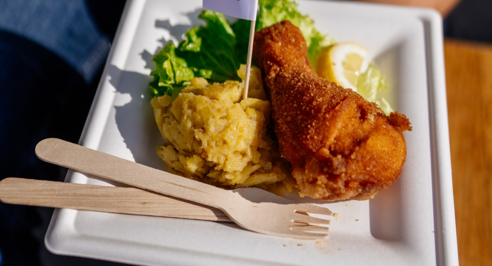 Ein weißer Pappteller mit einer knusprig gebratenen Hähnchenkeule, einer Portion Röstkartoffeln, einem grünen Salatblatt, einer Zitronenspalte sowie Holzbesteck und einem kleinen Fähnchen mit Logo.