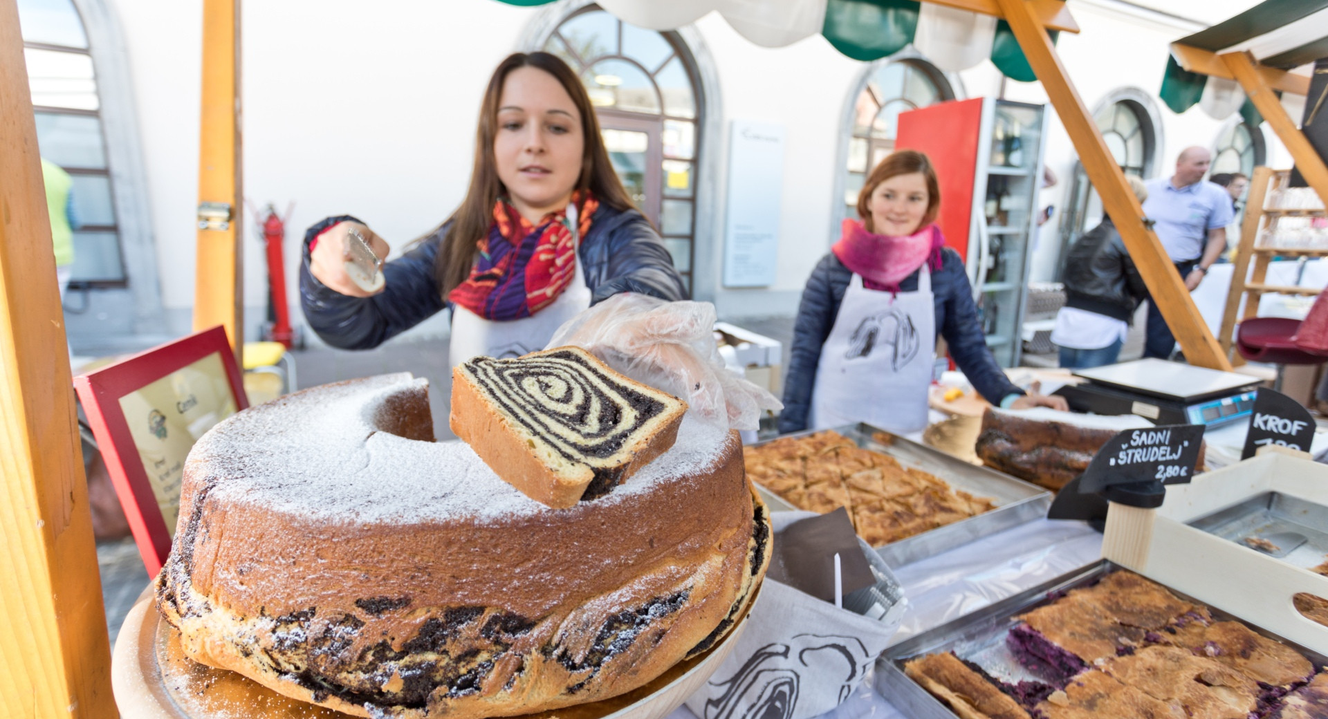 Eine Verkäuferin auf dem Markt Odprta kuhna schneidet eine frisch gebackene slowenische Mohnpotize an, die mit Puderzucker bestäubt ist.