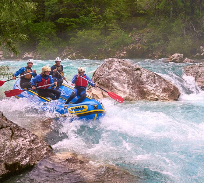 Un gruppo di quattro rafter su una barca lotta attraverso le rapide spumeggianti di un fiume turchese tra grandi rocce e una foresta verde.