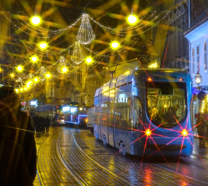 Il tram attraversa la città nel crepuscolo, con luci natalizie gialle che brillano sopra di esso.