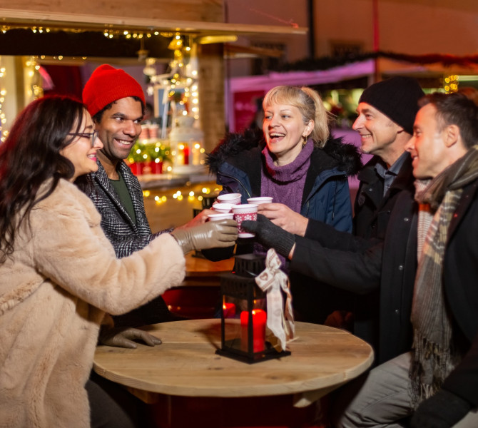 Cinque persone ben vestite stanno intorno a un tavolo rotondo, accanto a un banco, all'aperto, e brindano.