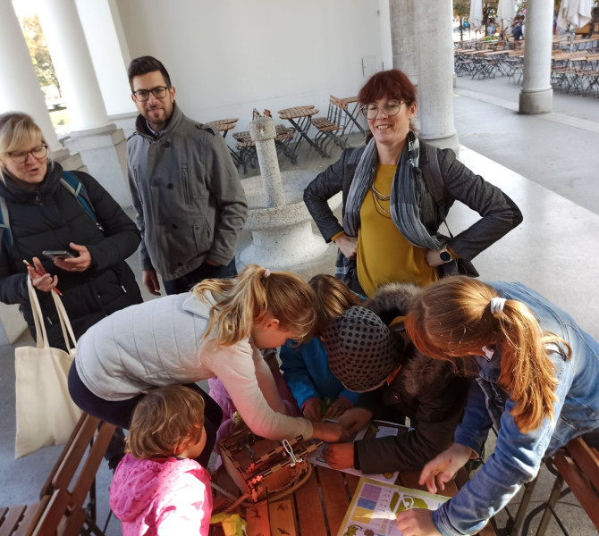 Starši opazujejo otroke, ki resujejo nalogo za mizo na ljubljanski trznici. 