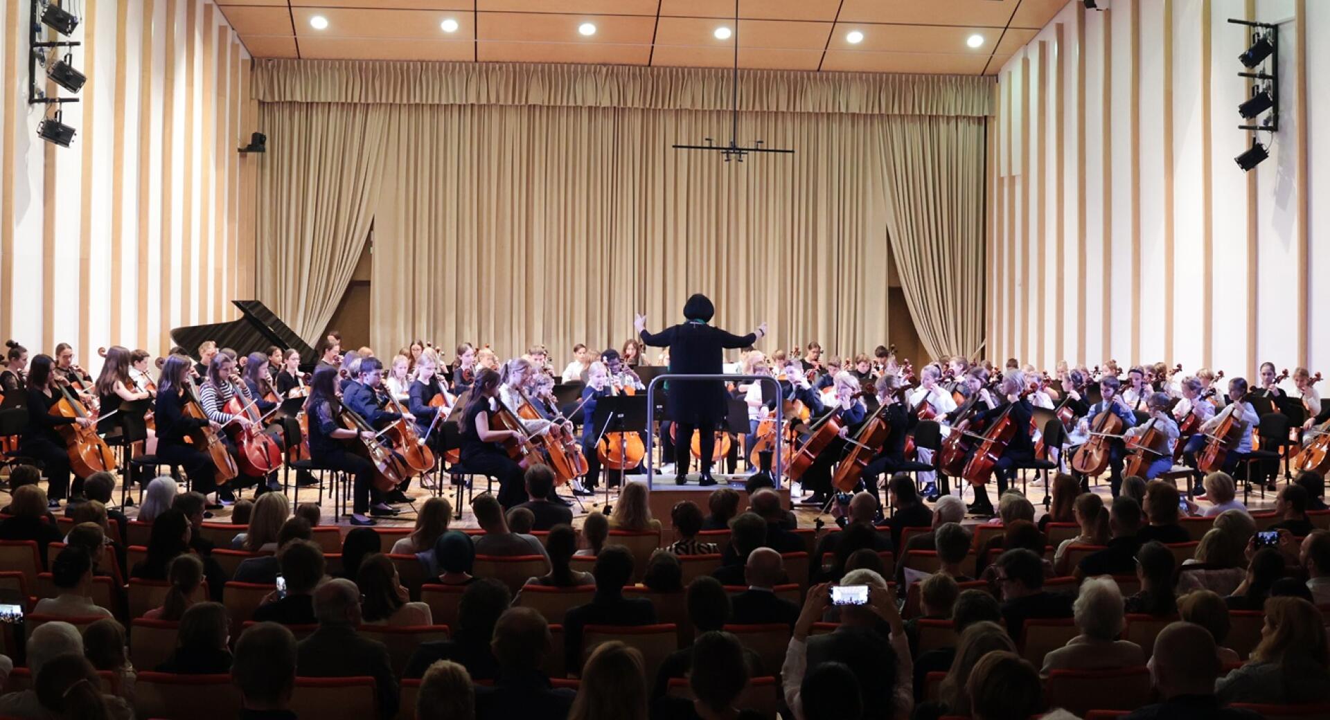 Dirigent vodi veliko skupino mladih violončelistov na odru v koncertni dvorani pred občinstvom.