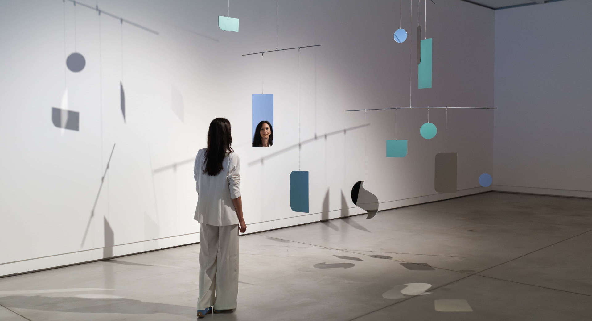 A woman in a white trouser suit stands in a white room, where mirrors of various shapes and sizes hang from the ceiling on strings at different heights.