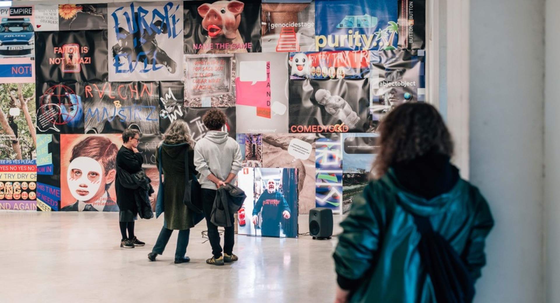 People are viewing an exhibition in a large hall with white floors, and on the wall there is a collage of graphics and photographs.
