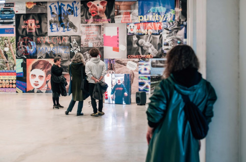 People are viewing an exhibition in a large hall with white floors, and on the wall there is a collage of graphics and photographs.
