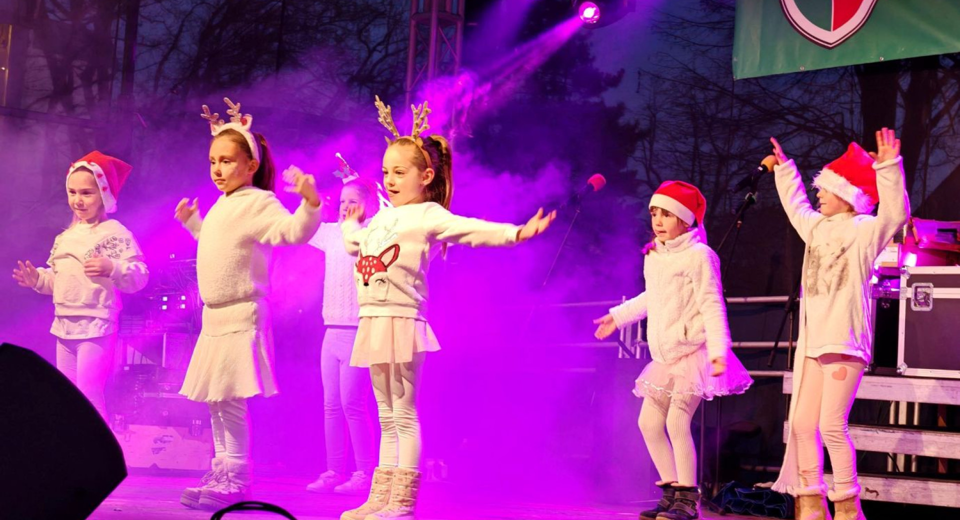 Otroci, obleceni v belo, z bozicnimi kapicami in jelenjimi rogovi, plesejo na odru.