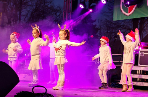 Otroci, obleceni v belo, z bozicnimi kapicami in jelenjimi rogovi, plesejo na odru.
