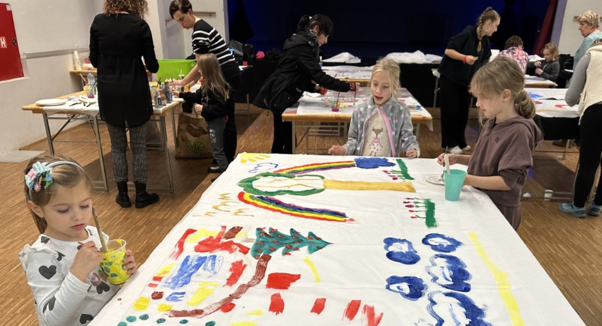 Na mizi je veliko belo platno, na katerega tri majhne deklice slikajo z raznobarvnimi temperami. V ozadju so se tri podobne mize, okoli katerih so ljudje in otroci.