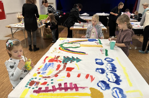 Na mizi je veliko belo platno, na katerega tri majhne deklice slikajo z raznobarvnimi temperami. V ozadju so se tri podobne mize, okoli katerih so ljudje in otroci.