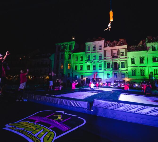 A night event in Ljubljana with trampolines set up in front of illuminated buildings. A person is seen performing a jump, suspended in mid-air.