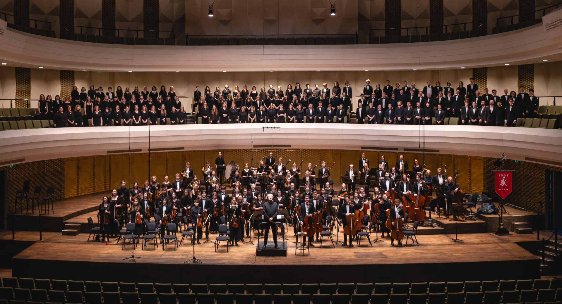 View of a large orchestra on stage with the audience on the balcony above.