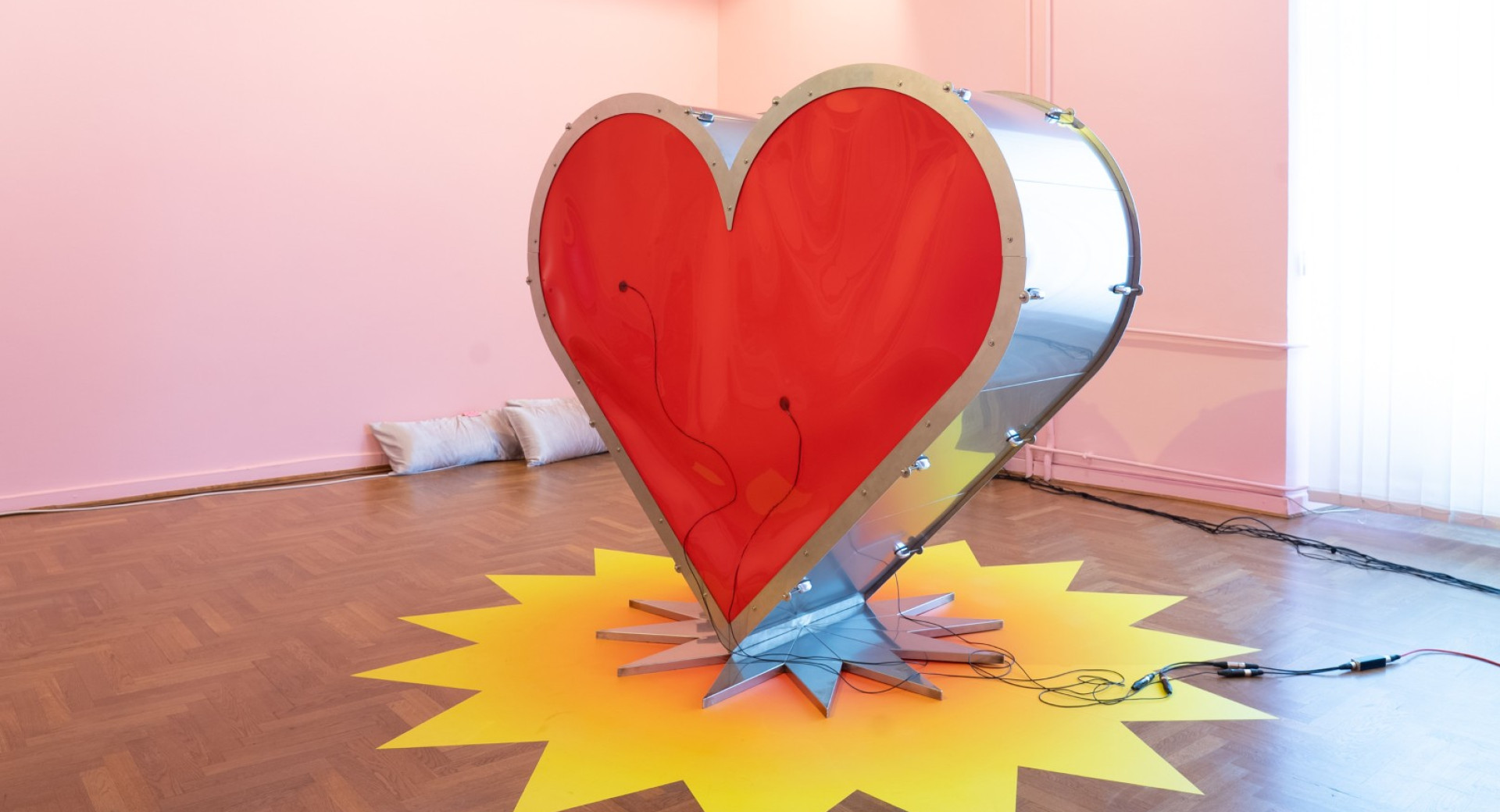 A large steel sculpture of a heart with a red centre, placed in a room on a yellow sun-shaped base.
