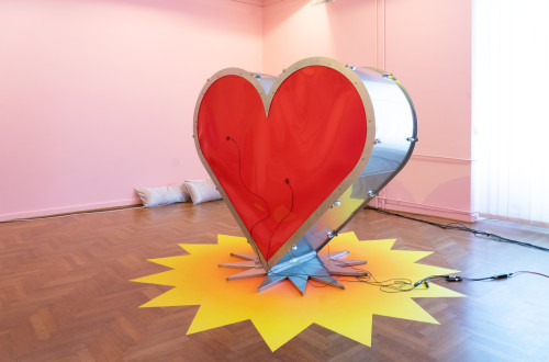 A large steel sculpture of a heart with a red centre, placed in a room on a yellow sun-shaped base.