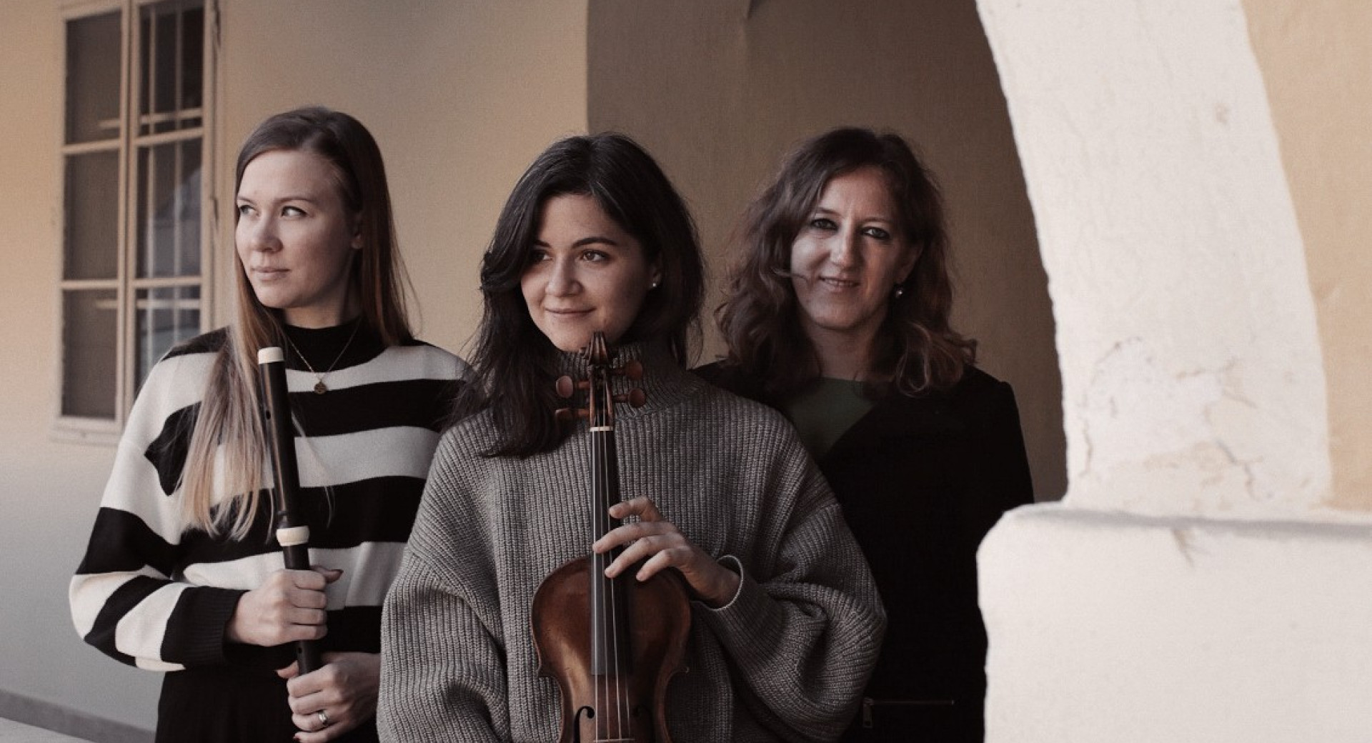Three women holding instruments are standing under a stone arch.