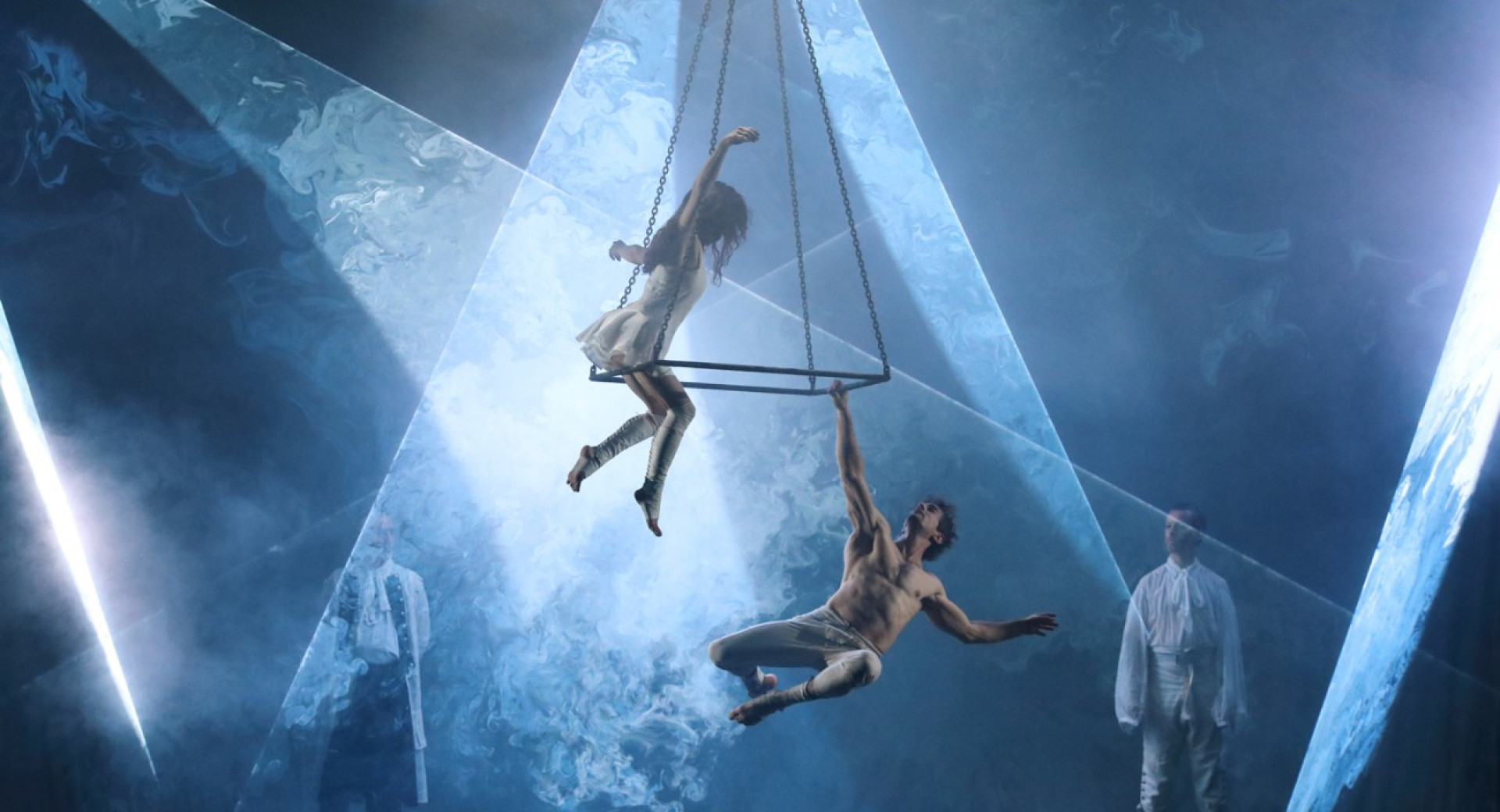 Akrobat in akrobatka v belih dresih izvajata akrobatski trik na odru na jekleni piramidi, ki visi s stropa.