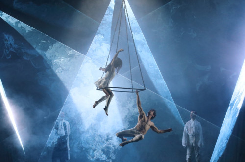 Akrobat in akrobatka v belih dresih izvajata akrobatski trik na odru na jekleni piramidi, ki visi s stropa.