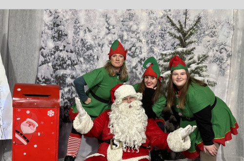 Tri skratinje stojijo okoli sedecega Bozicka in vsi skupaj pozirajo za fotografijo pred platnom z zasnezenimi smrekami.