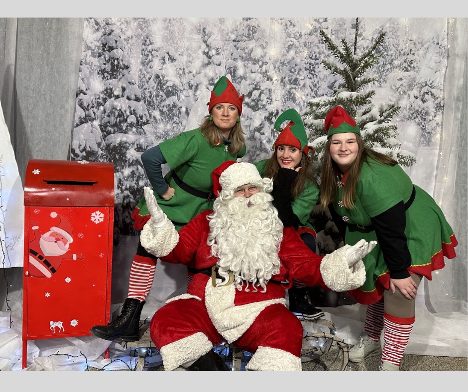 Tri skratinje stojijo okoli sedecega Bozicka in vsi skupaj pozirajo za fotografijo pred platnom z zasnezenimi smrekami.