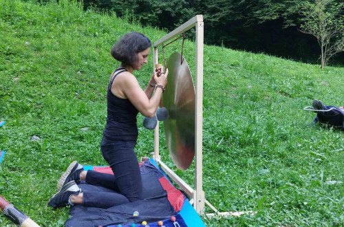 Zenska kleci na odeji na travi, pred seboj ima gong in druge pripomocke za zvocno meditacijo.