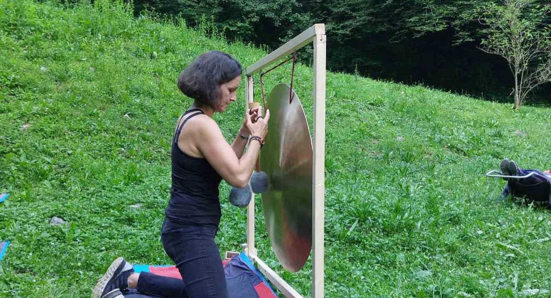 Zenska kleci na odeji na travi, pred seboj ima gong in druge pripomocke za zvocno meditacijo.
