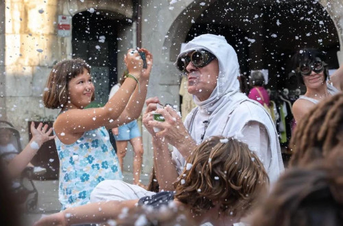 Moski v belem kostumu s kapuco nastopa na ulici, kjer pada umeten sneg, poleg njega pa deklica fotografira.