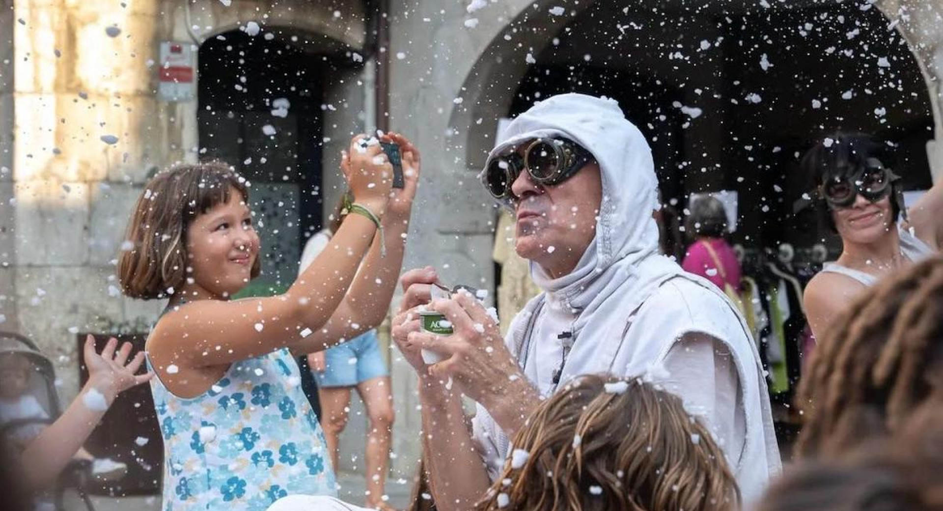 A man in a white costume with a hood is performing on the street as artificial snow falls, while a girl beside him is taking photos.