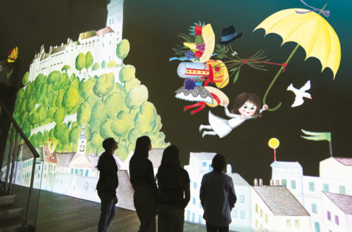 Stirje ljudje stojijo v senci pred veliko osvetljeno ilustracijo in jo opazujejo.