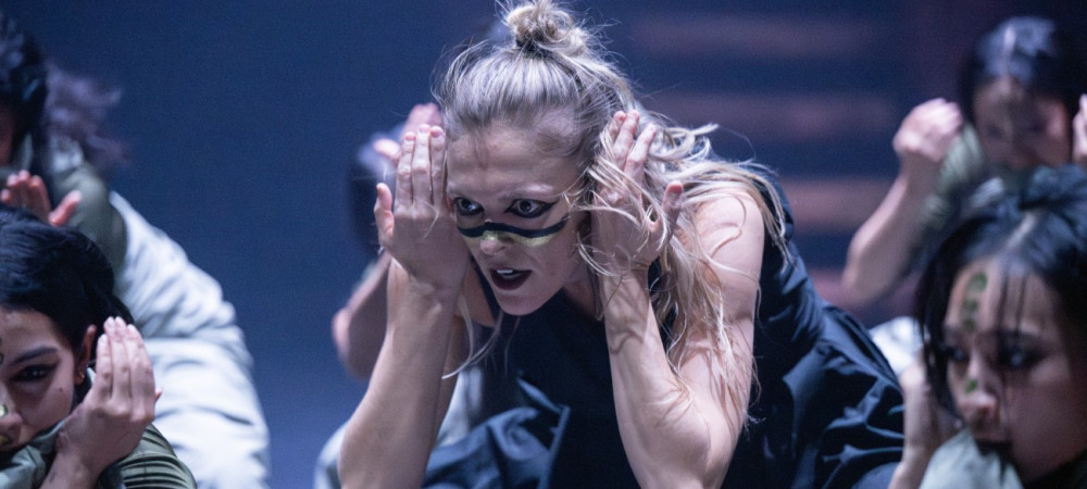 Bliznji posnetek zenske s plavimi lasmi, ki nastopa na odru v crni obleki in s poslikanim obrazom, v plesni pozi z rokami pri usesih.