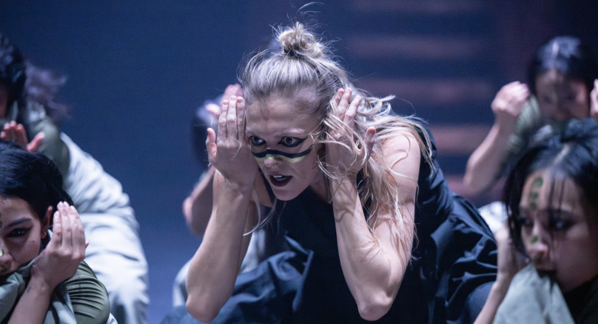 Bliznji posnetek zenske s plavimi lasmi, ki nastopa na odru v crni obleki in s poslikanim obrazom, v plesni pozi z rokami pri usesih.