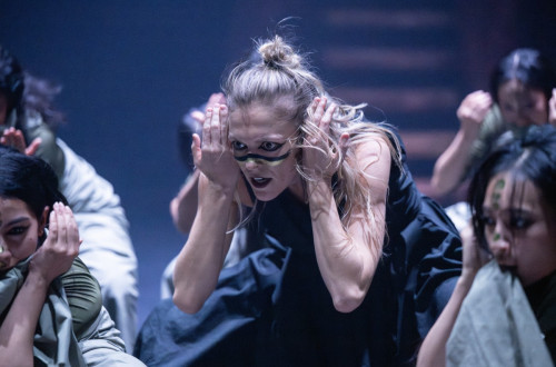 Bliznji posnetek zenske s plavimi lasmi, ki nastopa na odru v crni obleki in s poslikanim obrazom, v plesni pozi z rokami pri usesih.