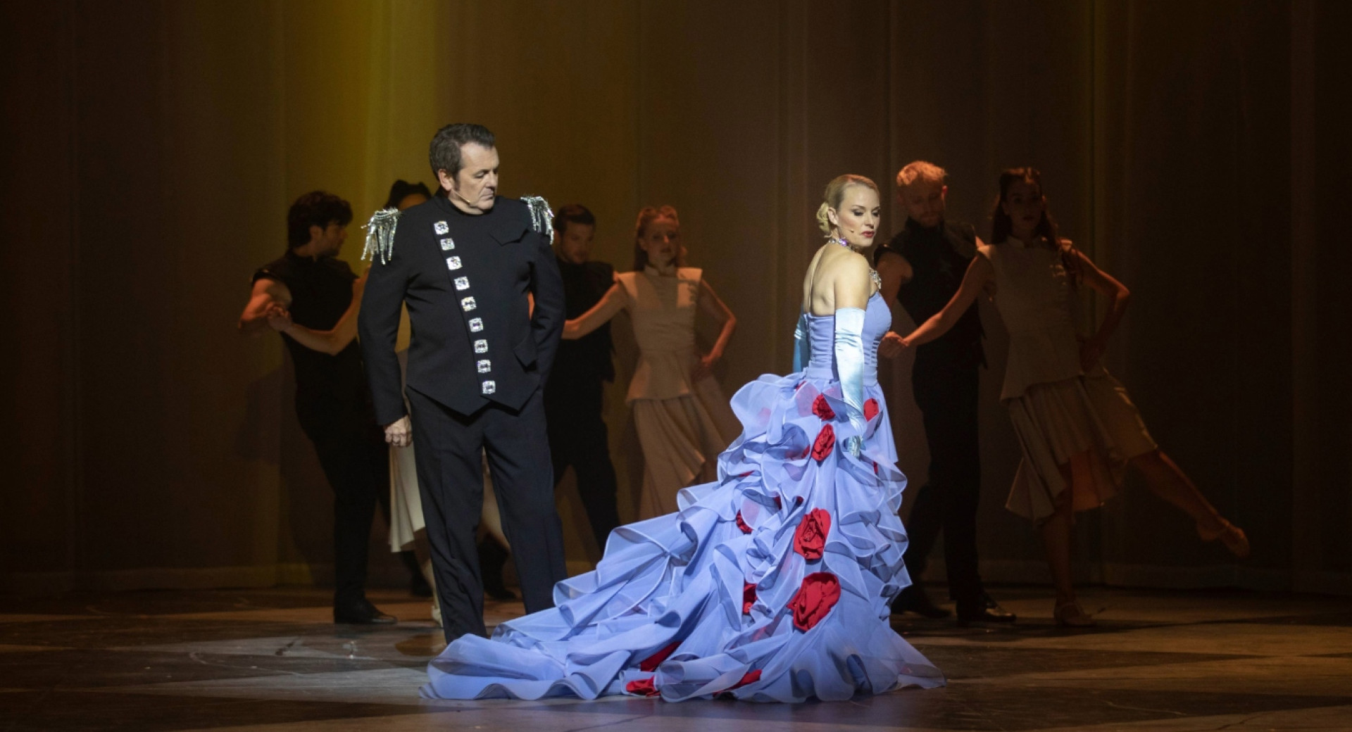 Svetlolasa zenska v dolgi svetlo modri obleki in moski v crni obleki na odru.