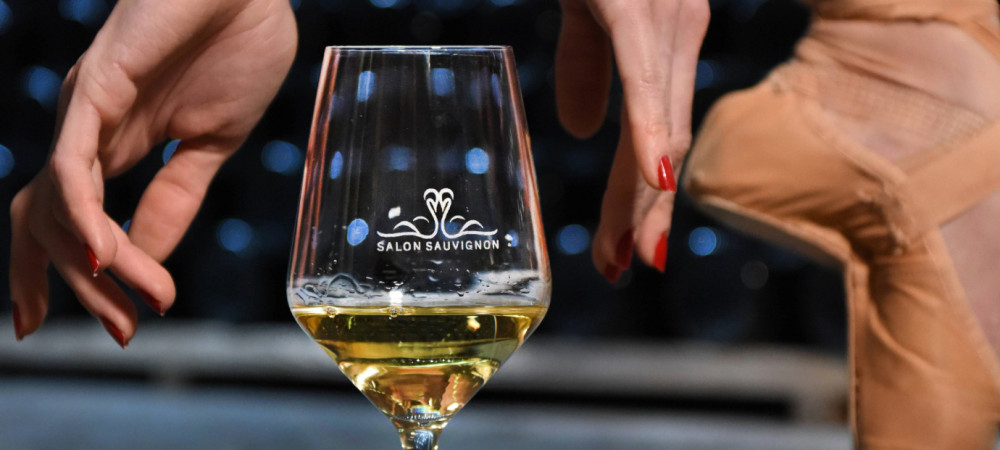 A glass of white wine next to a ballerina’s leg.