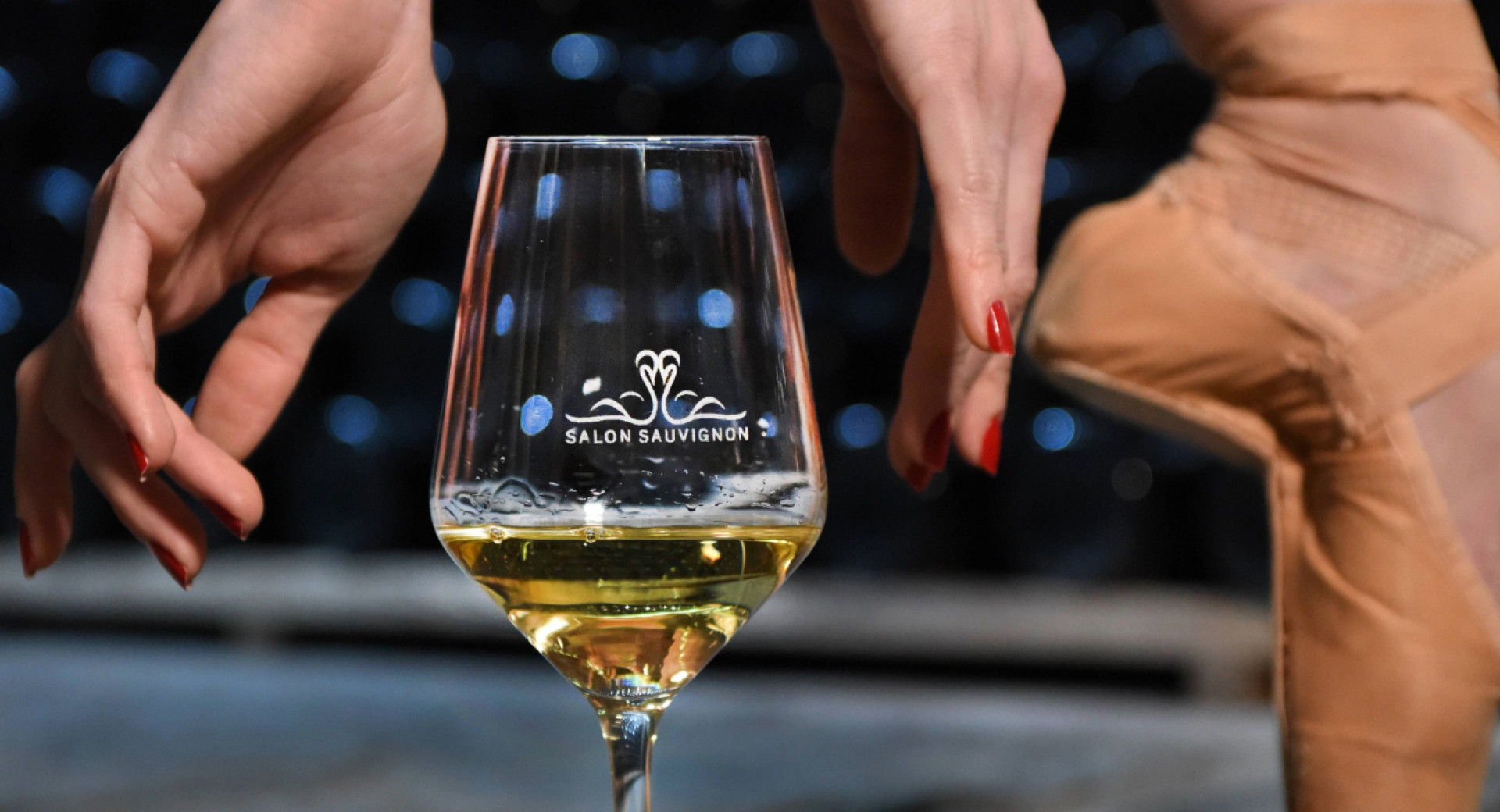A glass of white wine next to a ballerina’s leg.